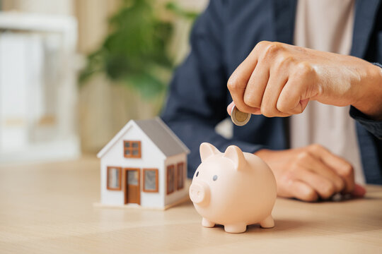 A businessman hand places a coin into a piggy bank beside a house model, symbolizing property savings, financial planning, and investment strategies for home buying