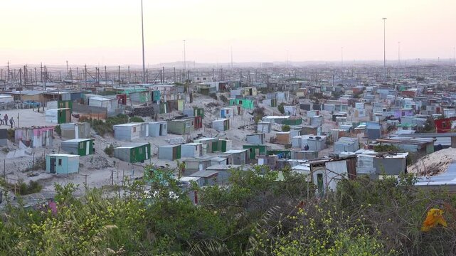 Wide angle view of extensive informal settlement with makeshift houses and corrugated metal shelters under a hazy sky in South Africa.