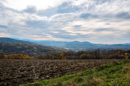 Expansive autumn panorama across cultivated farmland, wooded hills and scattered villages in the Beskid foothills of Lesser Poland. Layered mountain ridges under dramatic clouds create a strong landsc