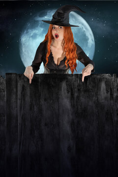 Red-haired witch in black hat peeking from behind dark wooden fence with empty copy space against the backdrop of the moon and night sky. Halloween holiday mockup for text or advertising design