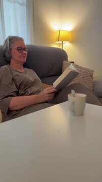 A woman sits on a couch in a living room reading a book. A warm lamp lights the space. A cup is placed on the table in front of her.