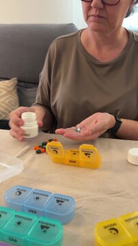 A person sits at a table arranging daily medication into colorful containers. The process takes place in a bright room with a couch in the background.