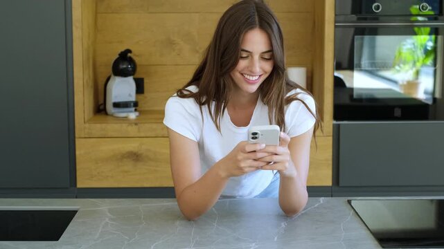 Beautiful happy woman leaning on her kitchen counter, laughing and smiling while using her mobile phone, enjoying online communication, funny content, or reading good news on social media