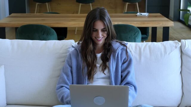 Smiling young woman sitting on a comfortable couch in her living room, typing on her laptop computer while working from home, chatting online, or studying for an online course