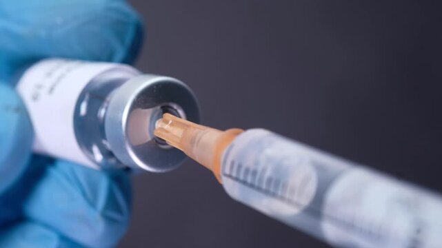 Gloved hand drawing covid-19 vaccine from glass vial into syringe close-up