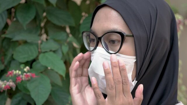 Side view of a modern Asian Muslim woman wearing a black hijab, cat eye glasses, and a protective white face mask outdoors.