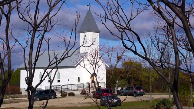 Exterior of a charming white country church with a steeple under a cloudy sky framed by bare tree branches.