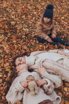 Mother kissing older daughter while lying on autumn leaves while younger daughter sits nearby with sad, lonely expression. Sibling rivalry, parent-child relationships, loneliness, exclusion, jealousy.