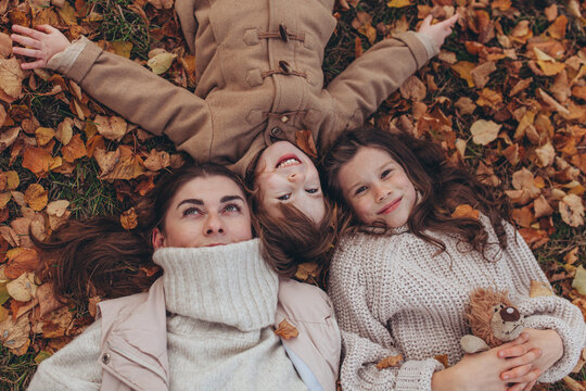 Top-down shot of happy mother and two children lying on ground, surrounded by colorful fallen autumn leaves. Family warmth, love, joy of autumn season, cozy seasonal fashion, mother's day.