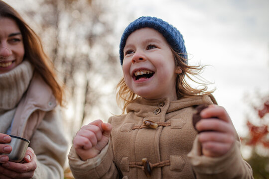 Joyful young girl wearing blue knitted hat and beige coat laughing while eating cookie outdoors, accompanied by her mother. Family happiness, carefree lifestyle, simple pleasures.