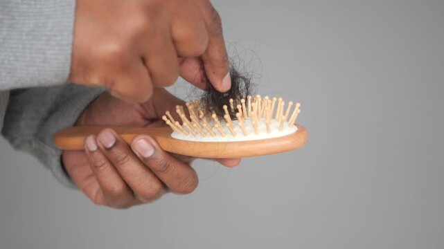 African man hand removing clump of fallen hair from wooden brush against gray background