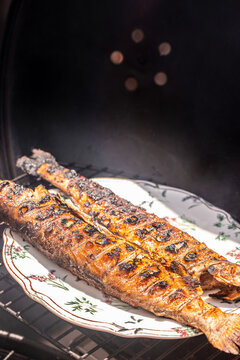 Grilled fish fillets on a decorative plate inside a barbecue grill, showcasing charred skin and grill marks, with smoke rising in the background