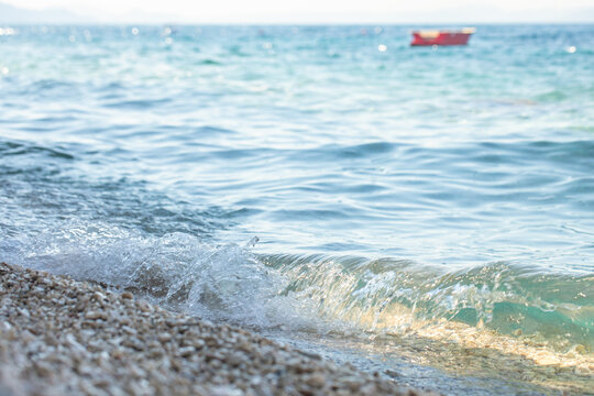 Gentle waves lapping against a pebbled beach with a distant red boat floating on the clear blue water under a bright sky