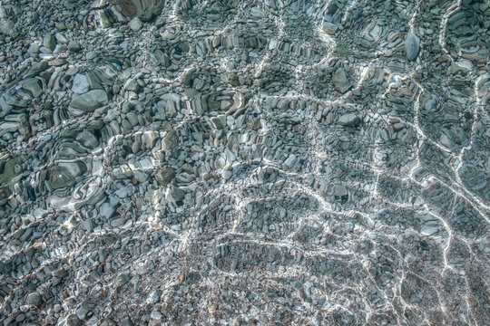 Clear water surface reflecting light over a bed of smooth pebbles and stones in a natural aquatic environment, showcasing intricate patterns and textures
