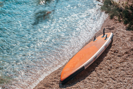 Orange stand-up paddleboard resting on a pebbled beach beside clear turquoise water under sunlight with gentle waves lapping at the shore