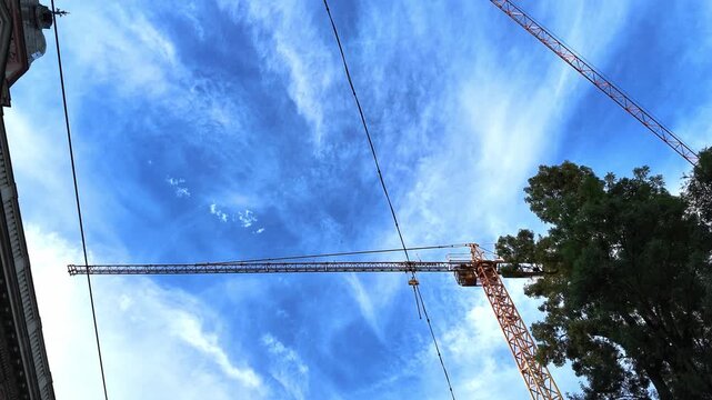 video looking upward at large tower cranes above an urban construction site, framed against a vivid blue sky with delicate high clouds and mature trees