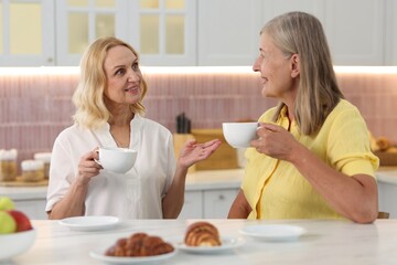Happy senior women with cups of drink spending time together at white table in kitchen