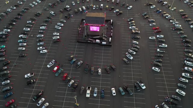 Aerial view of drive in concert stage in large parking lot at sunset showing circular car arrangement near ocean coast.