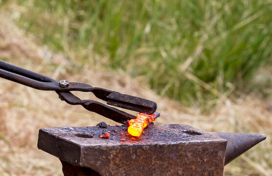 Close view of blacksmith tongs holding a heated orange iron rod on a weathered anvil, capturing traditional metalworking, forging heat, and handcraft outdoors