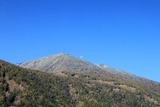 Rugged Mountain Peak with Moon Above Autumn Forest Slopes and Clear Blue Sky in Altay Mountains Kanas Xinjiang China