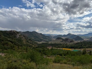 Fototapeta premium Forested Mountain Valley with Rocky Ridges and Scattered Buildings Beneath Dramatic Clouds
