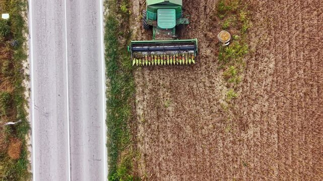 A tractor is harvesting crops in a field next to a road. A blue vehicle drives on the road. It is daytime and the field is dry and brown.