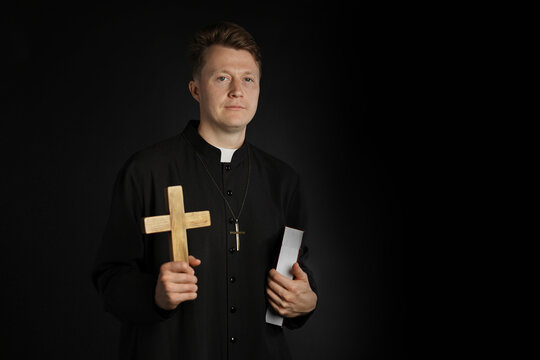 Priest in cassock with wooden cross and Holy Bible on black background, space for text