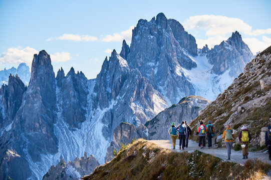 Hiking group follows exposed alpine road beneath towering snow-dusted Dolomites peaks, carrying backpacks and helmets after via ferrata adventure through dramatic high-altitude scenery in Italy