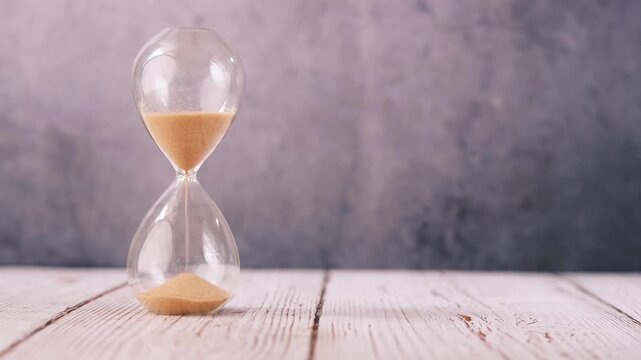 Clear hourglass showing sand falling on wooden table with grey background in motion, a symbol of passing time and urgency, a cinematic clip
