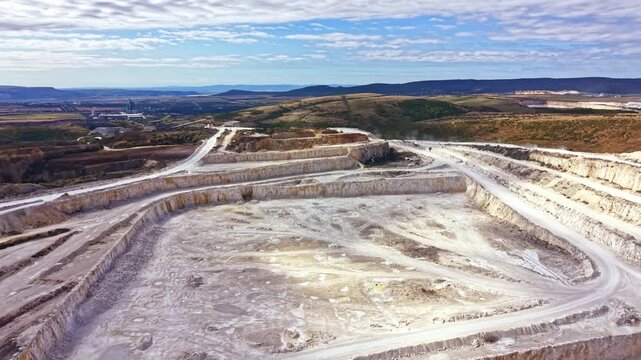 A mining site shows large areas of earth that have been excavated. Machines work on the ground, and mountains rise in the background. Clouds are visible in the sky.