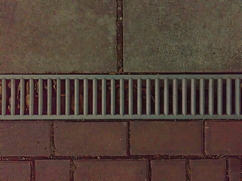 Top view of a metal drainage grate installed in a stone paved walkway. Urban infrastructure and stormwater management texture background.