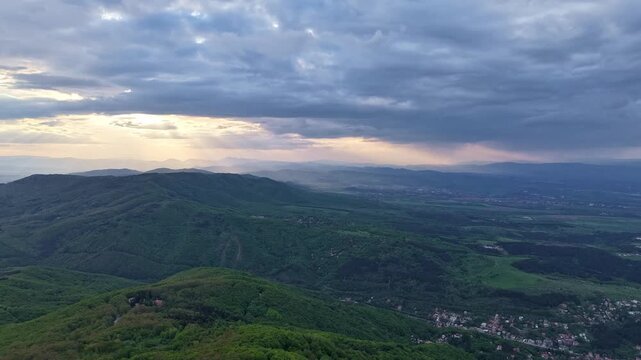 Layered forested mountains and scattered valley settlements fill the landscape during a slow aerial push-in beneath a cloudy evening sky.