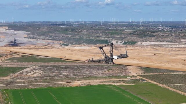 bucket wheel excavator works open pit lignite mine conveyor belts stretch across spoil heaps wind turbines line horizon green farm fields foreground kerpen germany hambach mine aerial view