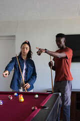 Fototapeta premium Diverse couple standing at red felt pool table, pointing and planning shots with cues, denim jacket