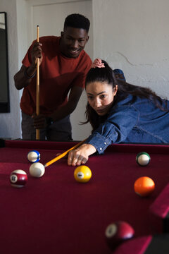 Diverse friends playing pool with woman leaning over burgundy felt table lining up shot with cues