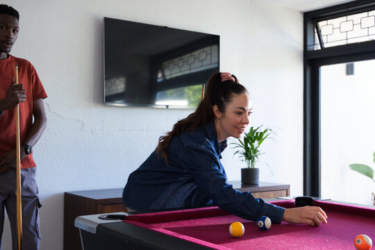 Diverse couple playing pool on burgundy-felt table in living room, female in denim jacket