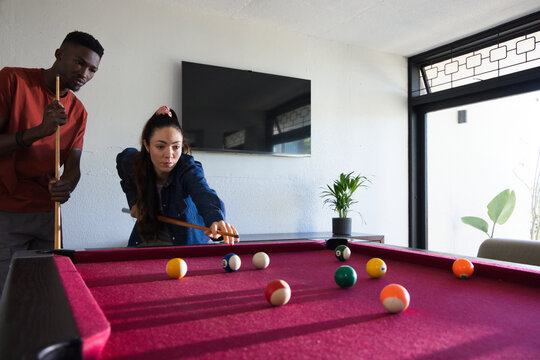 Diverse friends playing pool at home, woman lining up shot on magenta table, two cues, balls