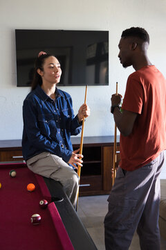 Couple holding wooden cues and chatting beside burgundy pool table with scattered balls at home