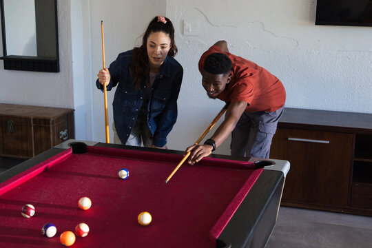 Diverse friends playing on red-felt pool table at home, woman wearing denim jacket, man aiming shot