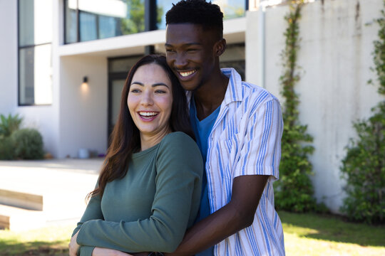 Diverse couple laughing and hugging, standing in front yard of modern house with large window