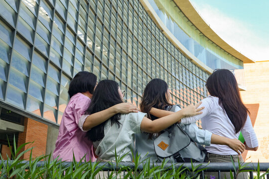 Rear view cheerful group Asian male and female students wearing backpacks arms around each other's shoulders in a display close friendship standing on the terrace in front university building.