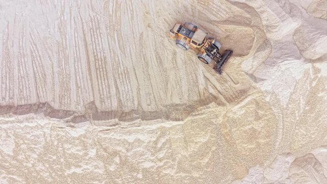 A large construction vehicle moves across an open field shaping the ground. Equipment impresses marks into the earth as it operates. Workers nearby observe the activity.