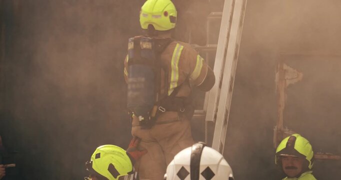 Firefighter crew &ndash; emergency service fireman rescue in action. In uniform and gas mask in response to fire with firefighting ladder against burning building releasing smoke.