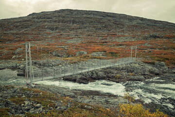 Enontekio, Finland - 31.08.2026: Metal suspension footbridge over rushing white water in rocky arctic tundra landscape under overcast sky © Andrei