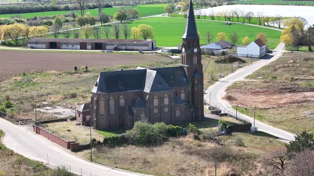 gothic revival church rises above plowed brown fields vivid green crops small pond glints beyond farm outbuildings scattered houses line distant road spire clock tower dominate skyline drone footage