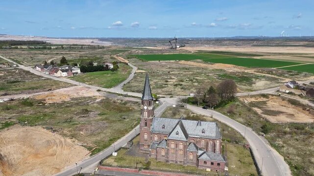 drone reveals brick church spire then pulls back across cleared demolition lots toward vast open pit mine bucket wheel excavator dwarfs farmland patchwork sandy overburden terraces stretch horizon