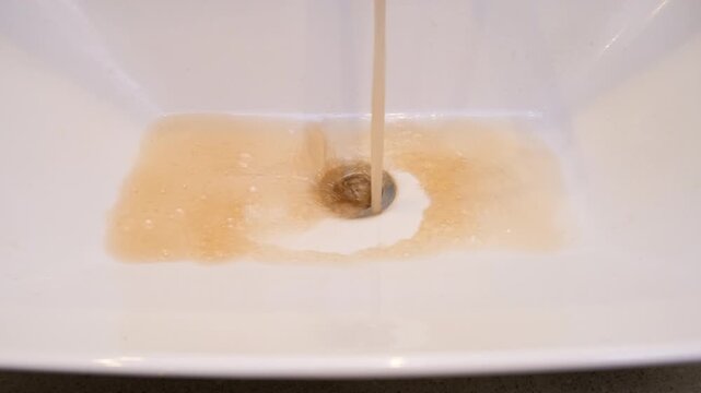 Discolored water from faucet flows into a white sink. The brown-orange liquid creates bubbles around the drain. This image highlights issues with water quality and plumbing.