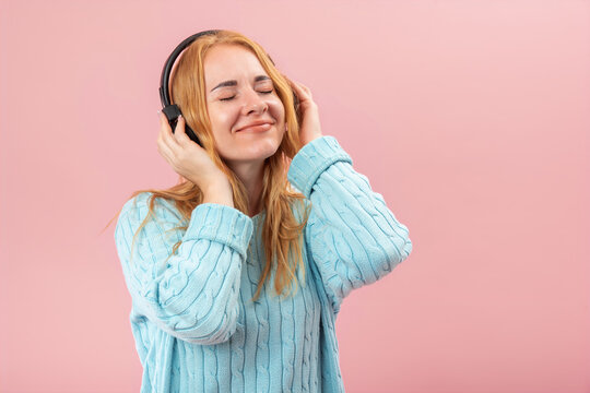 Enjoying favorite tunes, a blonde woman wearing a light blue sweater and black headphones is smiling with closed eyes, feeling happy and relaxed listening to the audio