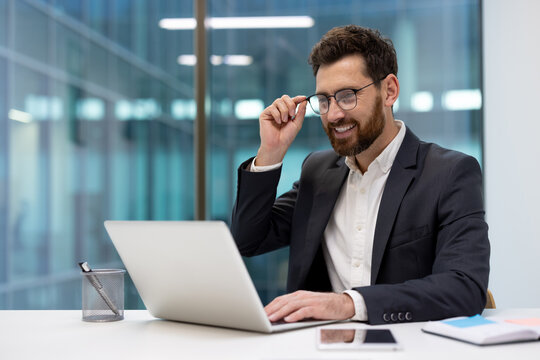 Smiling young businessman sitting at a desk in the office and talking on a video call on a laptop, holding glasses in his hand