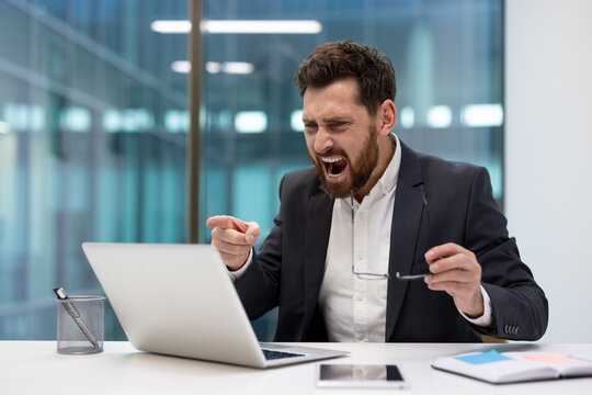 Angry young businessman sitting in the office at the table and talking on a video call on a laptop, holding glasses in his hand, shouting at the screen